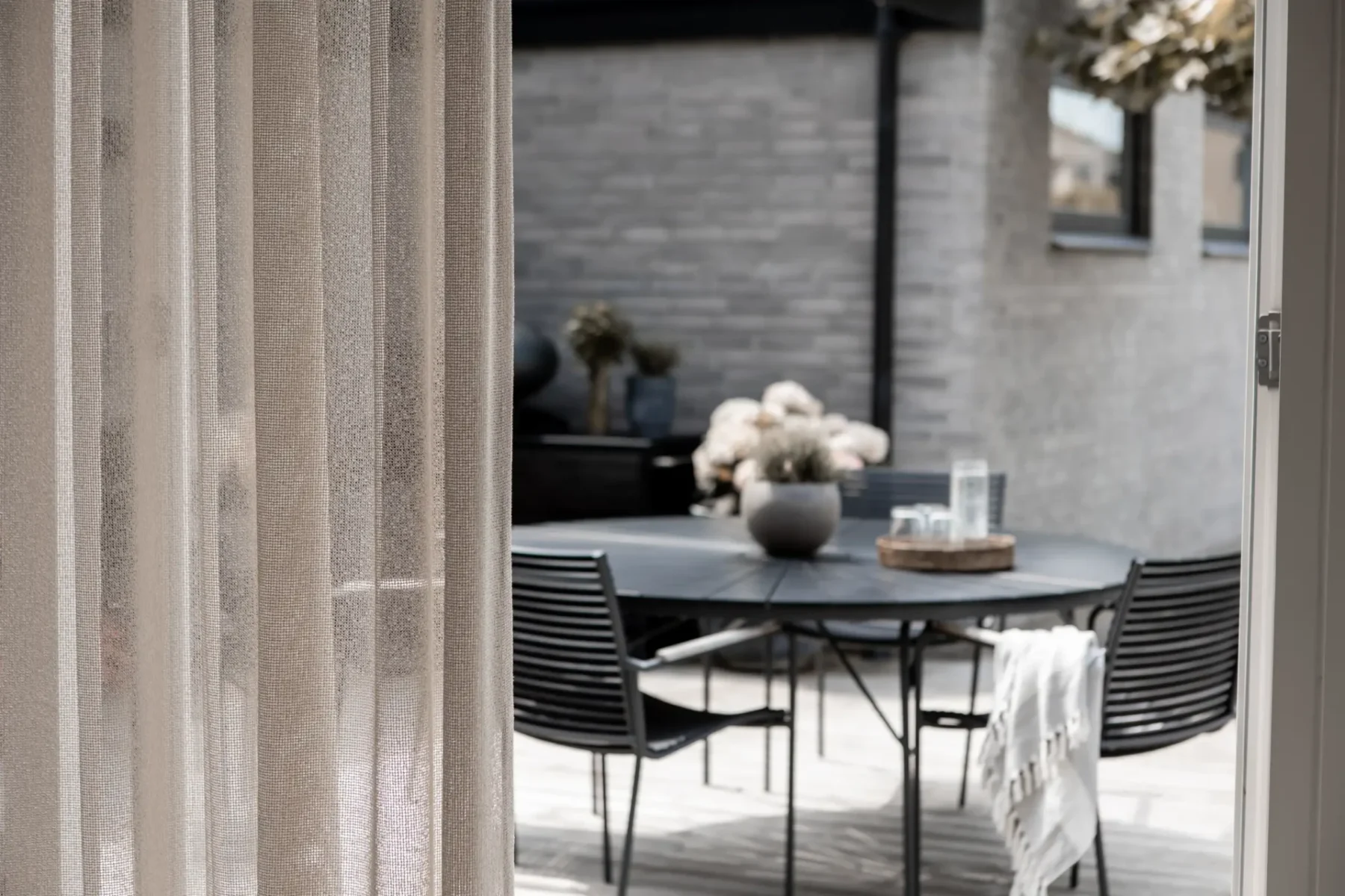 Närbild på en skir beige gardin med en uteplats med runt bord i bakgrunden