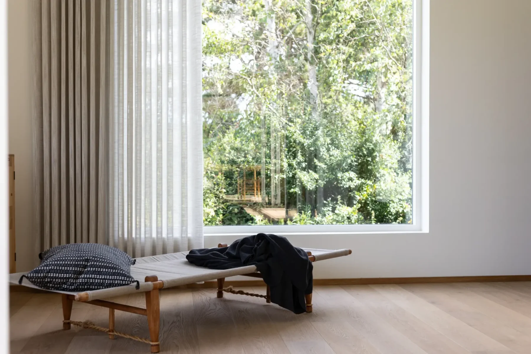 Skira beige gardiner vid ett stort fönster mot en trädgård, med en dagbädd framför