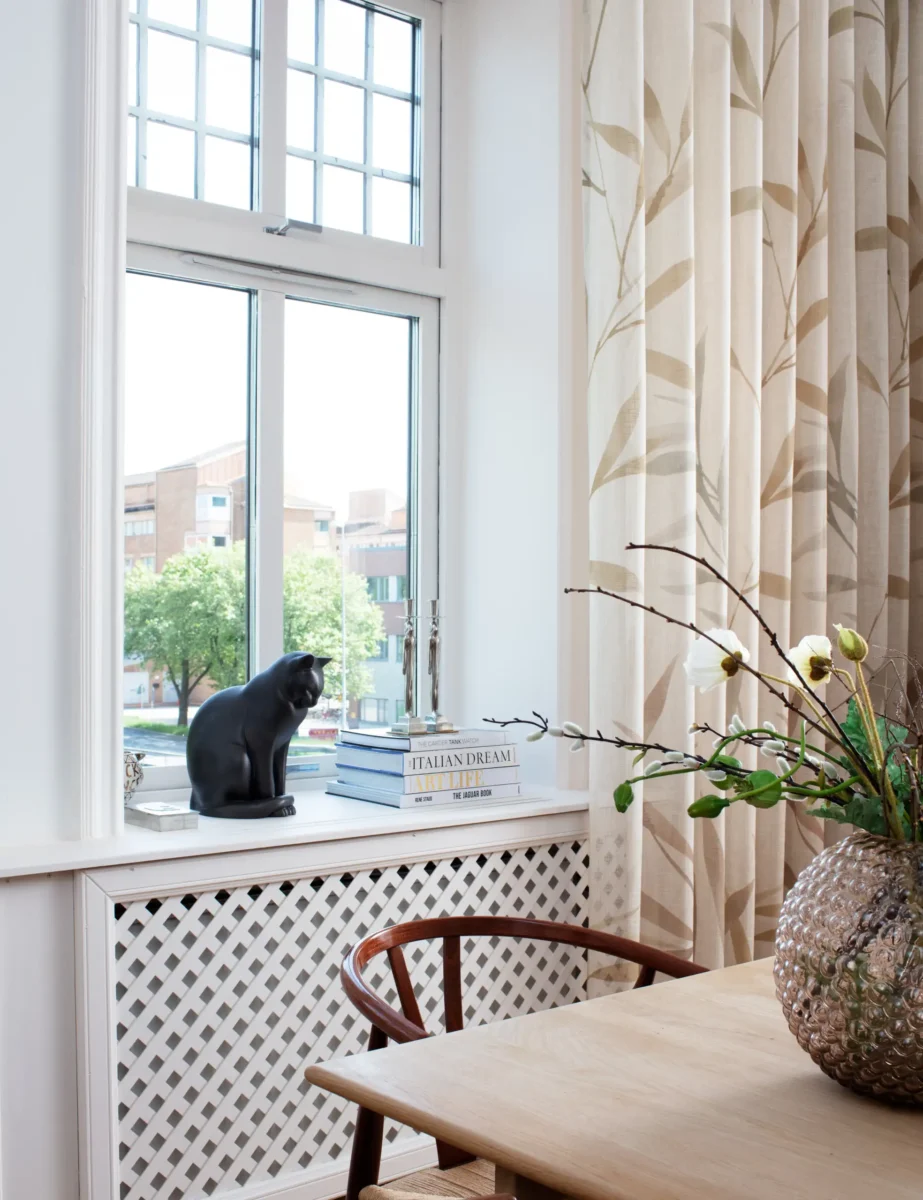 Skir gardin med bladmönster vid ett spröjsat fönster med en svart kattfigur i fönstret