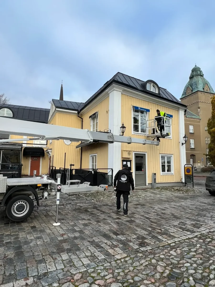 Montör i skylift arbetar vid ett fönster på ett gult hus medan en kollega går mot liftbilen