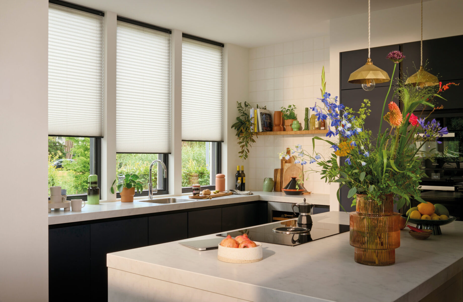 Vita plisségardiner i tre köksfönster bakom en köksö med en stor blombukett