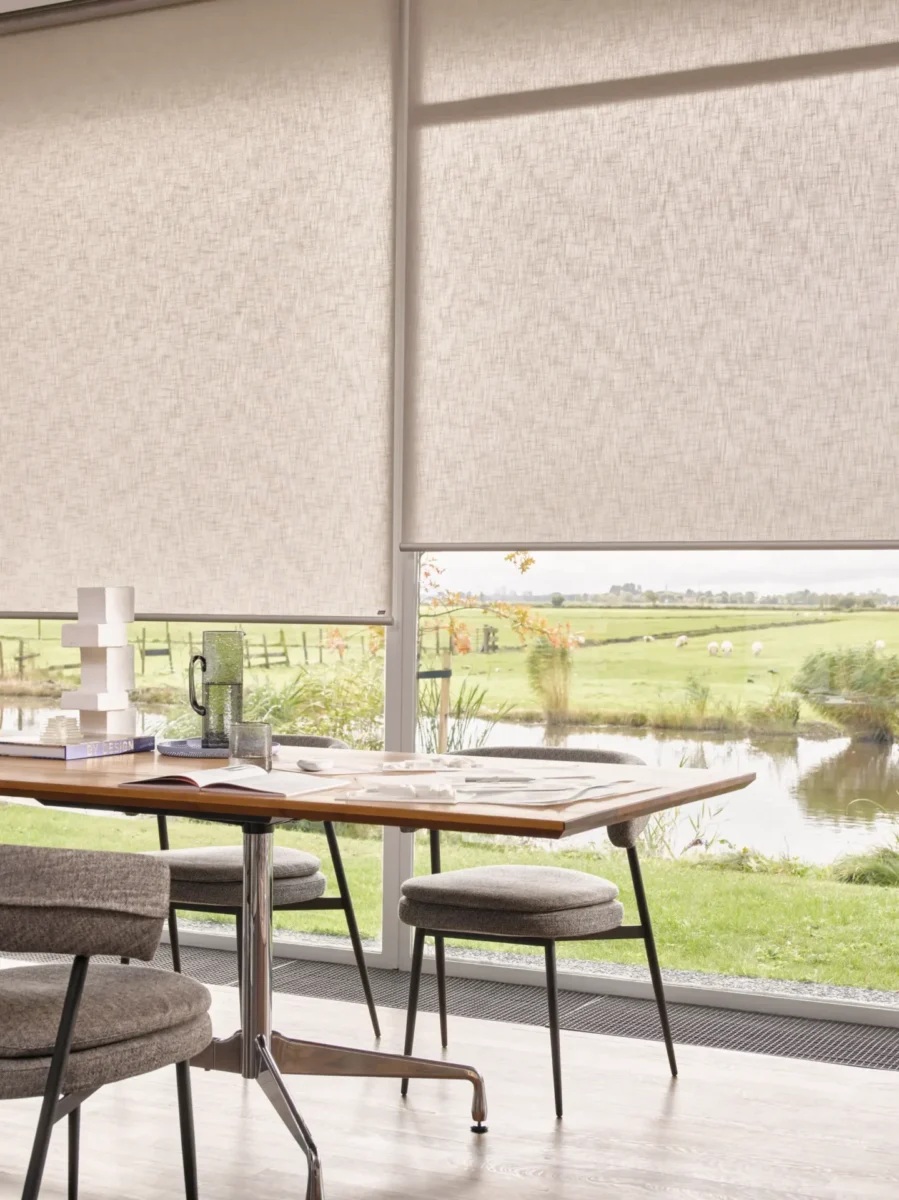 Beige rullgardiner halvt nerdragna i stora fönster med utsikt över ängar och vatten