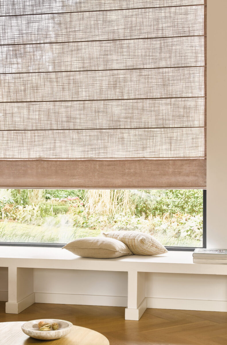 Beige hissgardin med brun nederkant i ett fönster ovanför en vit sittbänk